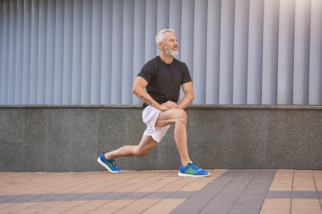 Active middle aged sportive man in sports clothing stretching his legs warming up and exercising