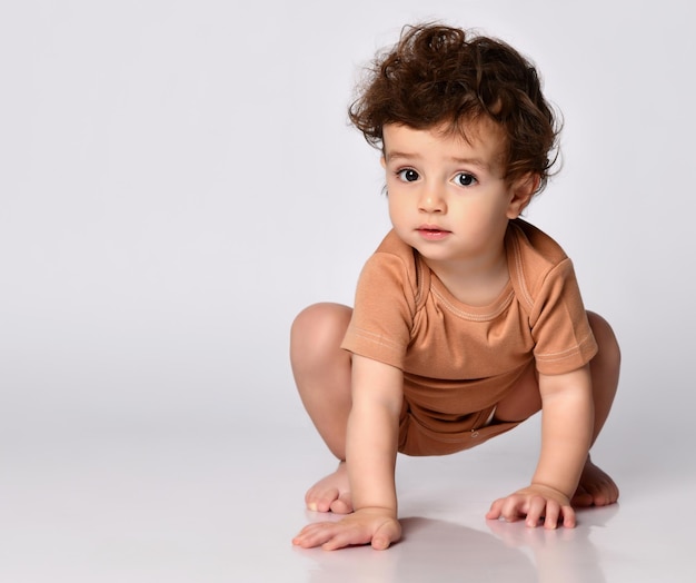 Active period in infants Charming darkhaired curly baby boy crawls on all fours on a gray background Child dressed in a brown bodysuit tries to get to his feet Place for text