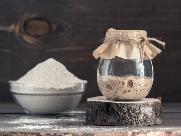 Active rye sourdough starter in glass jar and rye flour on brown wooden background starter for sourdough bread toned image copy space