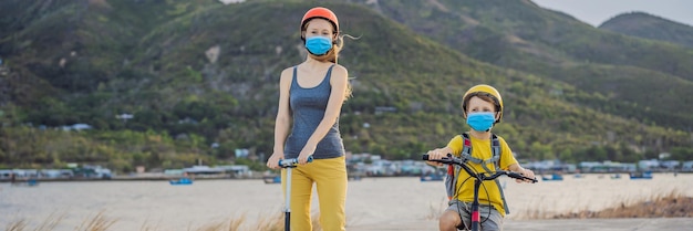 Active school kid boy and his mom in medical mask and safety helmet riding a bike with backpack on