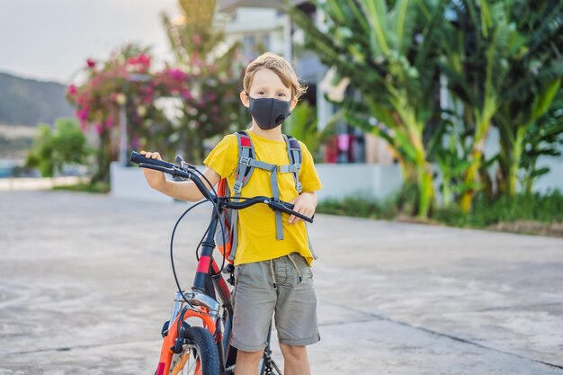 Active school kid boy in medical mask riding a bike with backpack on sunny day Happy child biking on way to school You need to go to school in a mask because of the coronavirus epidemic