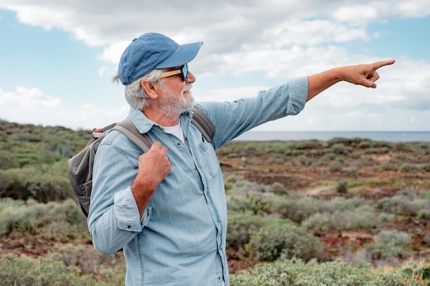 Active senior man in casual shirt and hat carries backpack walking in country footpath enjoying