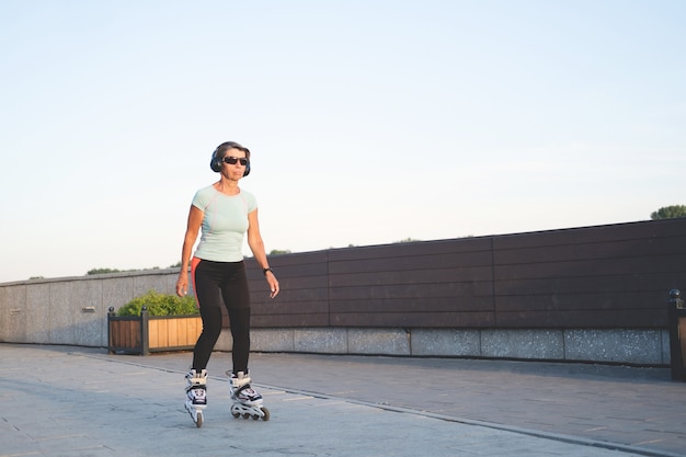 Active seniors caucasian woman roller blading in city outdoor. listen music. Headphones. Healthy lifestyle. Fitness. embankment. High quality photo