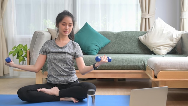 Active young woman in sportswear exercising with dumbbells and watching fitness lessons online on laptop