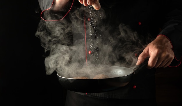 Adding salt to meat steaks in a hot pan with steam in the hands of a professional chef