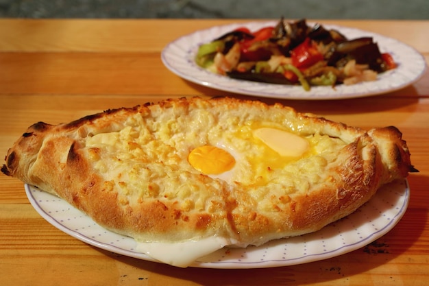 Adjaruli Khachapuri Traditional Georgian Cheese and Egg Bread with Eggplant Salad in Background