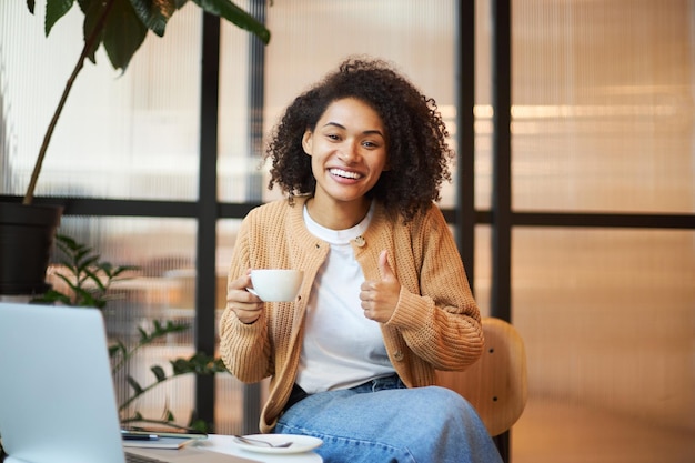 Adorable African American woman showing thumb up and smiling looking at camera while a coffee break