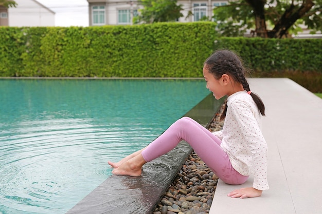 Adorable Asian young girl child having fun play water by her feet at poolside near the swimming pool