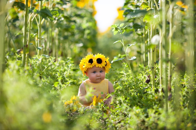 Adorable baby girl with a sunflower wreath sitting on the sunflowers field and investigated the nature around