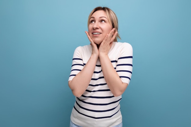 Adorable blond girl in a striped sweater is surprised to the side on a blue background with copy