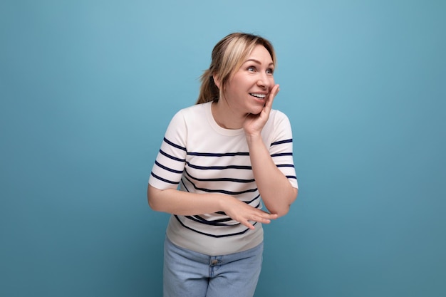 Adorable blond girl in a striped sweater tells a secret quietly on a blue background with copy space