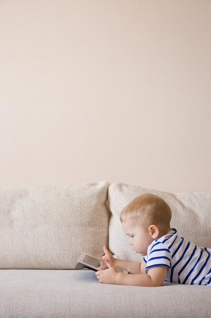 Adorable blond toddler boy laying on the sofa and playing with tablet pc at home indoors