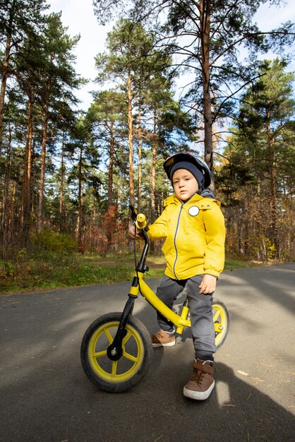 Adorable boy in casualwear and protective helmet standing by his balance bike on asphalt road against pinetrees in natural environment