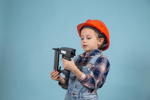 Adorable caucasian child is wearing orange safety helmet,holding construction stapler at hands. Concept of building a house for a happy family