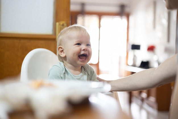 Adorable cheerful happy infant baby boy child smiling while sitting in high chair at the dining table in kitchen at home beeing spoon fed by his mother