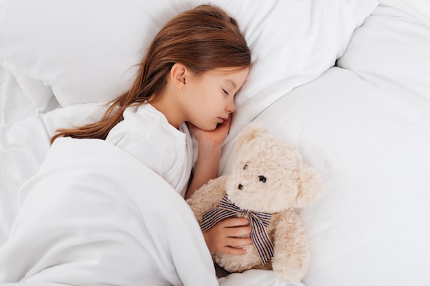 Adorable child resting on bed and keeping her hand on teddy bear while having pleasant dreams