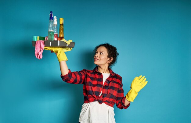 Adorable curly woman in yellow rubber gloves raising arms with a tray of cleaning supplies in hand and looking up on a blue