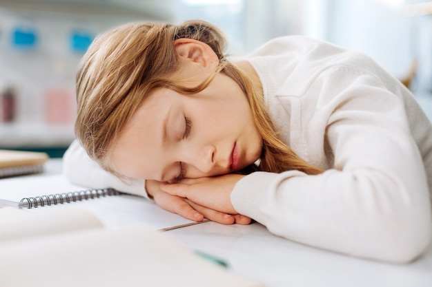 Adorable fair-haired little girl sleeping over her books while sitting at the table and doing homework