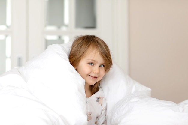 Adorable funny kid with blue eyes in pajamas lies in bed under a white fluffy blanket