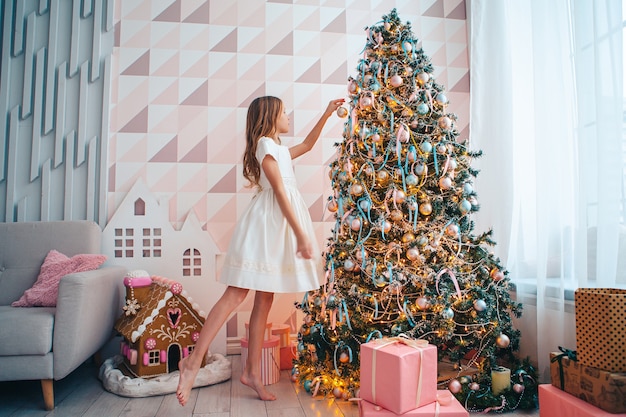 Adorable girl decorates the Christmas tree indoors. Happy Christmas holidays