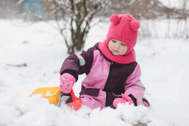 Adorable girl dig snow with shovel and pail on playground covered with snow little girl playing in winter outdoors