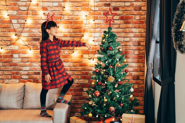 adorable girl kid in reindeer decorating christmas tree with baubles at home. little child standing high on sofa holding golden ball. lights hanging on red brick wall in background.