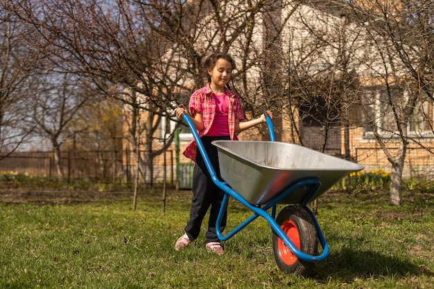 Adorable girl pushing wheelbarrow on a farm. Farming and gardening for small children. Outdoor summer activities for little kids.