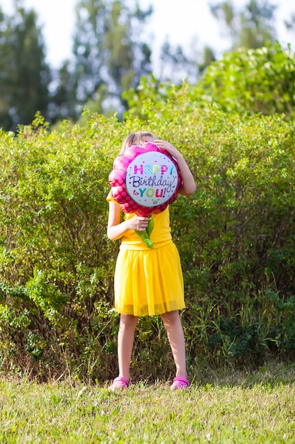 Adorable girl in yellow dress with festive happy birthday colorful balloon in the shape of flower. Happy child outdoors