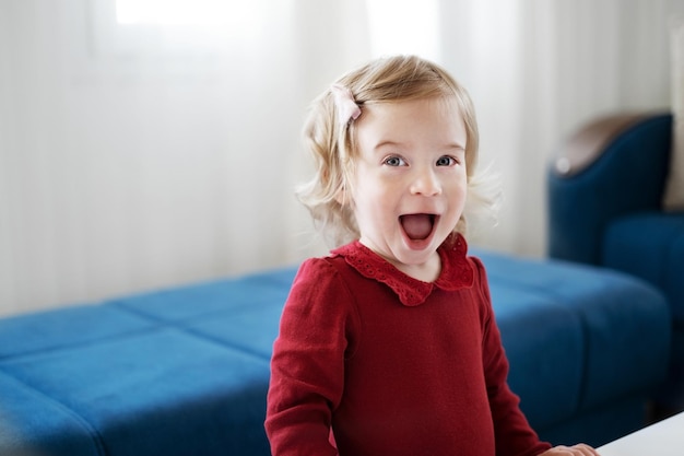 Adorable happy amazed open mouth caucasian blonde baby girl toddler infant smiling laughing looking at camera Pretty emotional child with curly hair having fun indoors near sofa