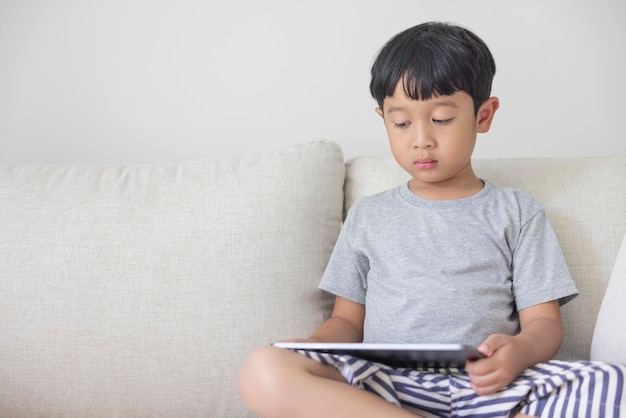 Adorable happy Asian boy wearing a gray shirt and bluewhite striped shorts is having fun playing with his tablet on a cream sofa looking at the mobile screen