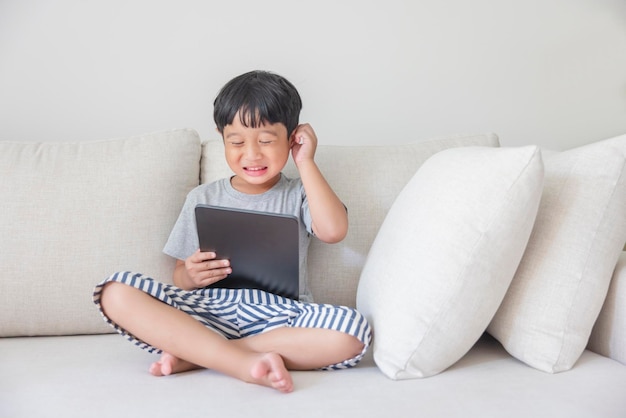 Adorable happy Asian boy wearing a gray shirt and bluewhite striped shorts is having fun playing with his tablet on a cream sofa looking at the mobile screen