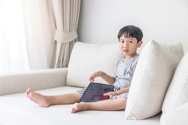 Adorable happy Asian boy wearing a gray shirt and bluewhite striped shorts is having fun playing with his tablet on a cream sofa looking at the mobile screen