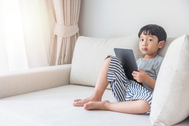 Adorable happy Asian boy wearing a gray shirt and bluewhite striped shorts is having fun playing with his tablet on a cream sofa looking at the mobile screen