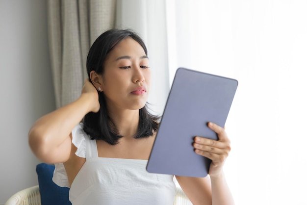 Adorable happy Asian woman wearing white dress is having fun playing with his tablet on a cream sofa looking at the mobile screen