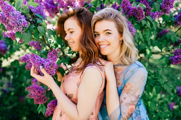 Adorable happy cheerful fabulous twin sisters in different beautiful summer dresses posing in spring blooming park.