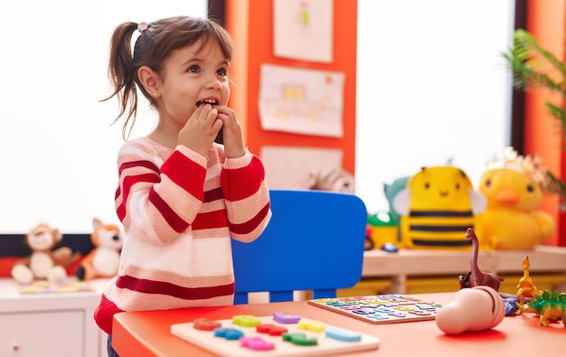 Adorable hispanic girl playing with maths puzzle game standing at kindergarten