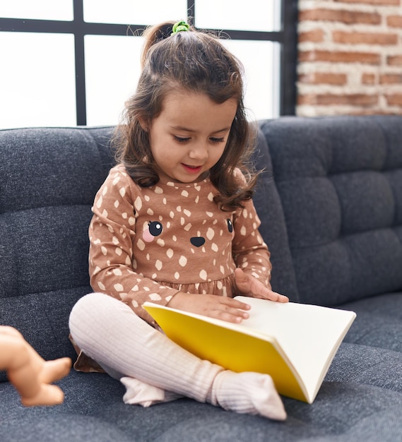 Adorable hispanic girl reading book sitting on sofa at home