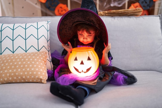 Adorable hispanic girl wearing halloween costume holding pumpkin basket lamp at home