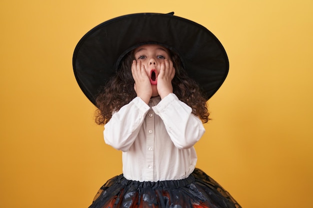 Adorable hispanic girl wearing halloween costume standing with surprise expression over isolated yellow background