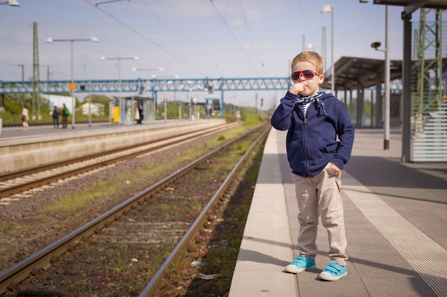 Adorable kid boy waiting for a train at the railway station in a sunny day Child ready for traveling by train Train station Dreaming of traveling Sunny day at the railway station