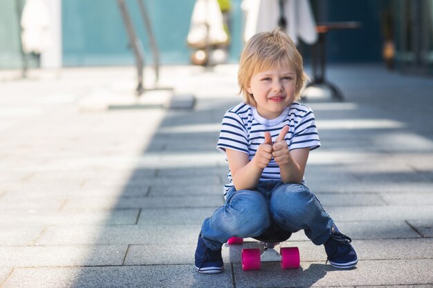 Adorable kid outdoors. Cute pretty child smiling at camera. Casual boy on summer time skating on a skateboard. thumb up