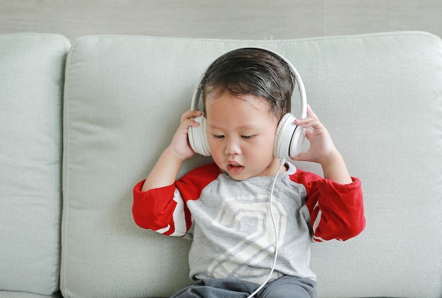 Adorable little Asian baby boy in headphones is using a smartphone lying on the sofa at home Child listening to music on earphones