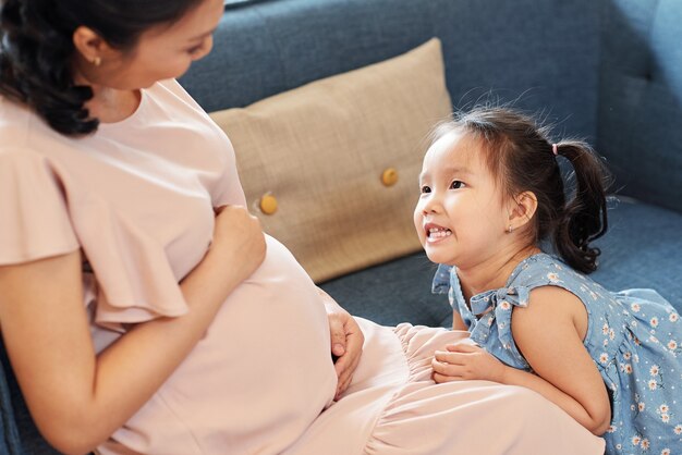 Adorable little Asian girl talking to her pregnant mother about sibling she is expecting