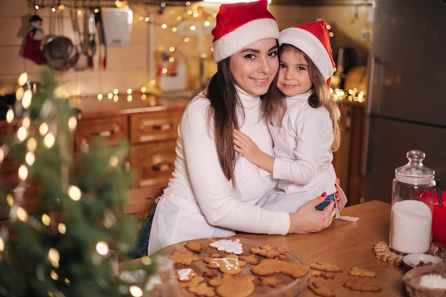 Adorable little daughter with her mom at kitchen during christmas holidays decorative fit tree at