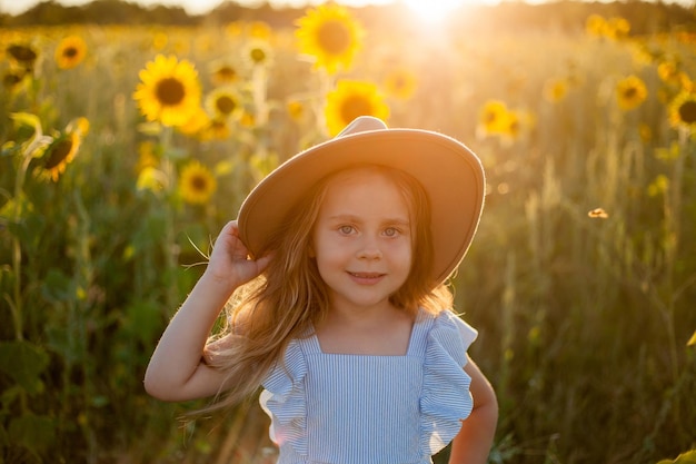 Adorable little girl 4 years old with curly long blond hair in a blue sundress and hat in a sunflower field Summer portrait of a happy child Sunset