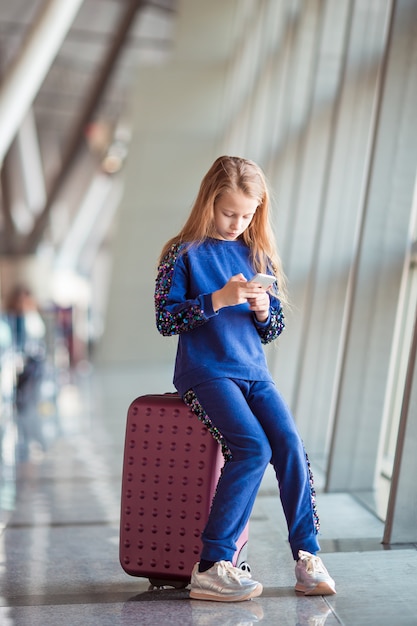 Photo adorable little girl in airport near big window playing with her phone