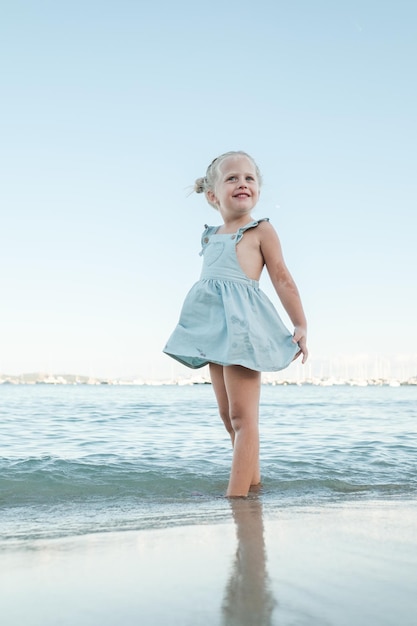 Adorable little girl in blue dress standing near waving sea and looking away against cloudless sky during summer vacation