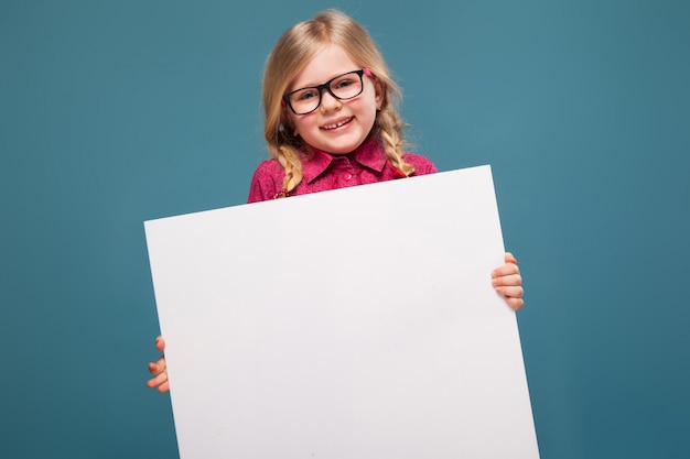 Adorable little girl in pink shirt, black trousers and glasses holds empty poster