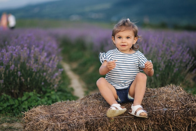 Adorable little girl sits on hay by the farm Background of summer lavender field Cute girl in striped tshirt and blue shorts