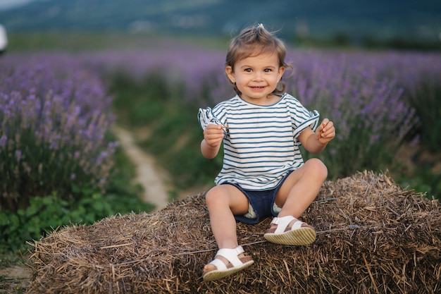 Adorable little girl sits on hay by the farm Background of summer lavender field Cute girl in striped tshirt and blue shorts